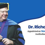 Dr. Richard Gayer, Chief Academic Officer of IAU, appointed as TRACS Commissioner and Institutional Representative; standing in academic regalia beside blue background featuring IAU logo and text announcing his leadership role with the Transnational Association of Christian Colleges and Schools.